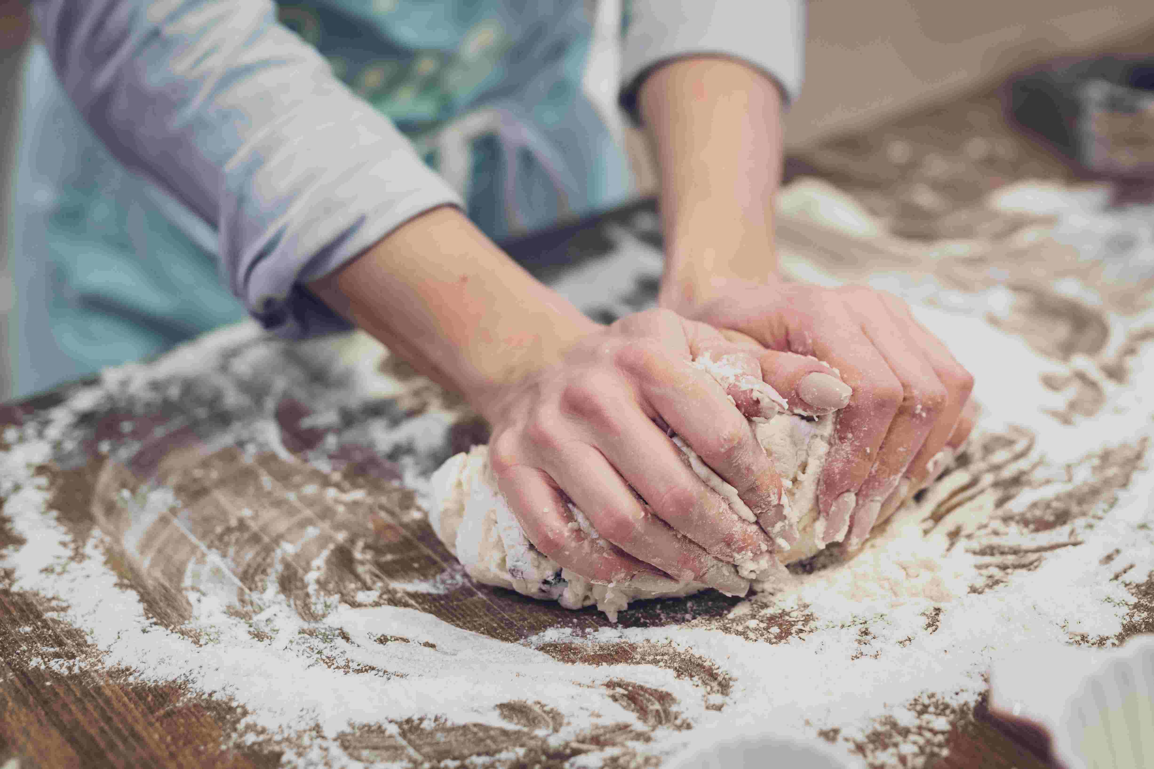 Womens hands kneading a dough on a kitchen island covered in flour.